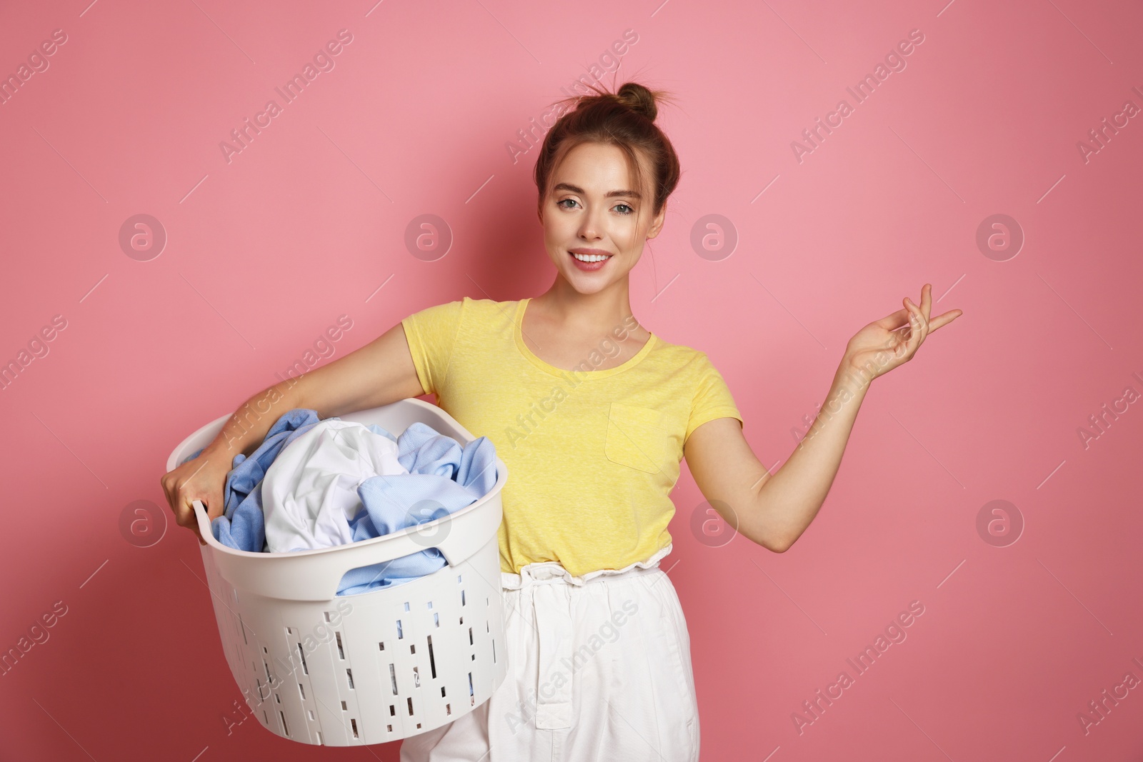 Photo of Happy young housewife with basket full of laundry on pale pink background