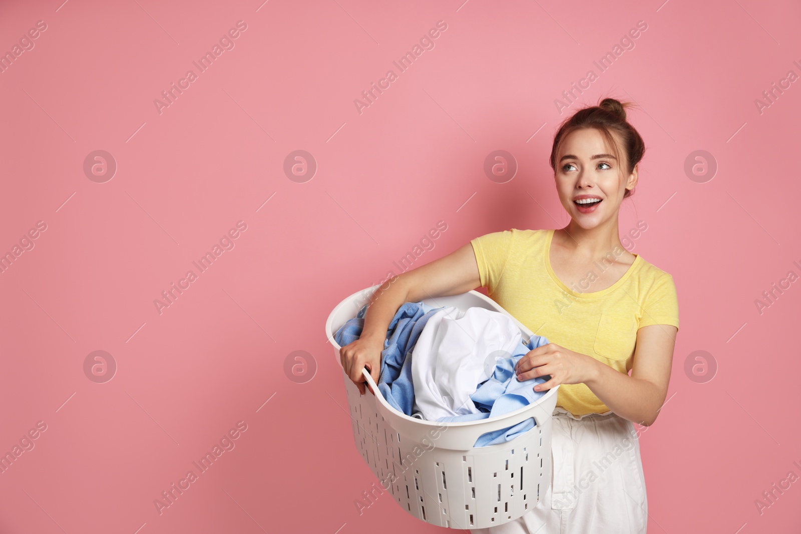 Photo of Happy young housewife with basket full of laundry on pale pink background. Space for text