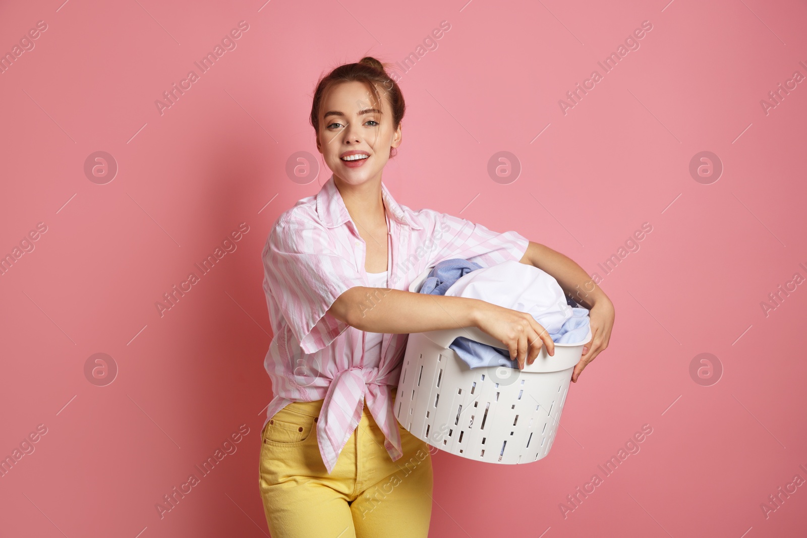 Photo of Happy young housewife with basket full of laundry on pale pink background