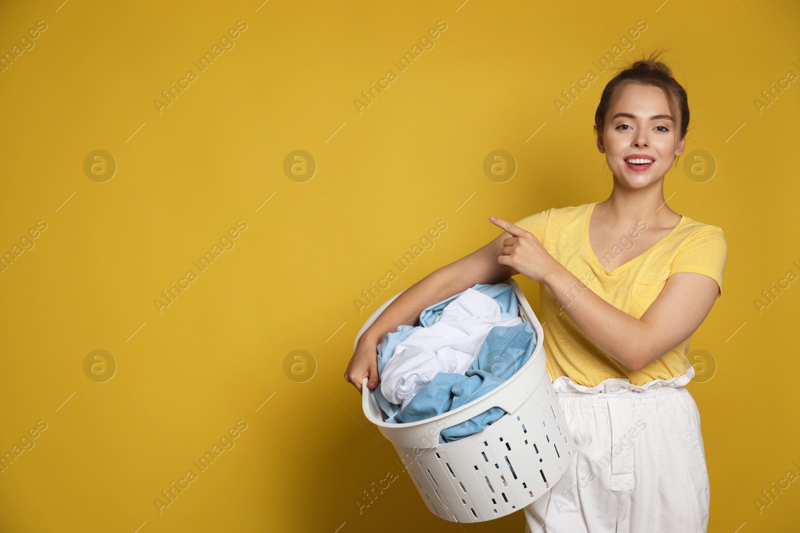 Photo of Happy young housewife with basket full of laundry on yellow background. Space for text