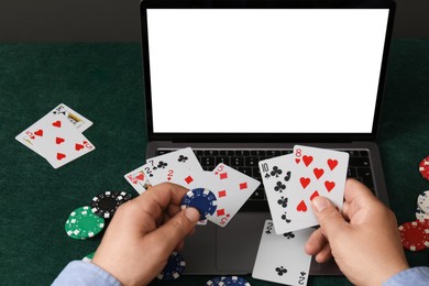 Photo of Online poker. Man holding playing cards and chip at green table with laptop, closeup