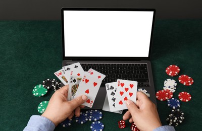 Photo of Online poker. Man holding playing cards at green table with laptop and chips, closeup