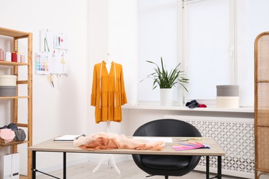 Photo of Different fashion designer`s supplies on wooden table and mannequin with new dress in workshop