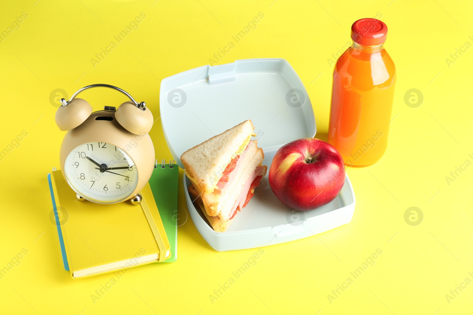 Photo of Lunch box with snacks, bottle of juice, alarm clock and books on yellow background