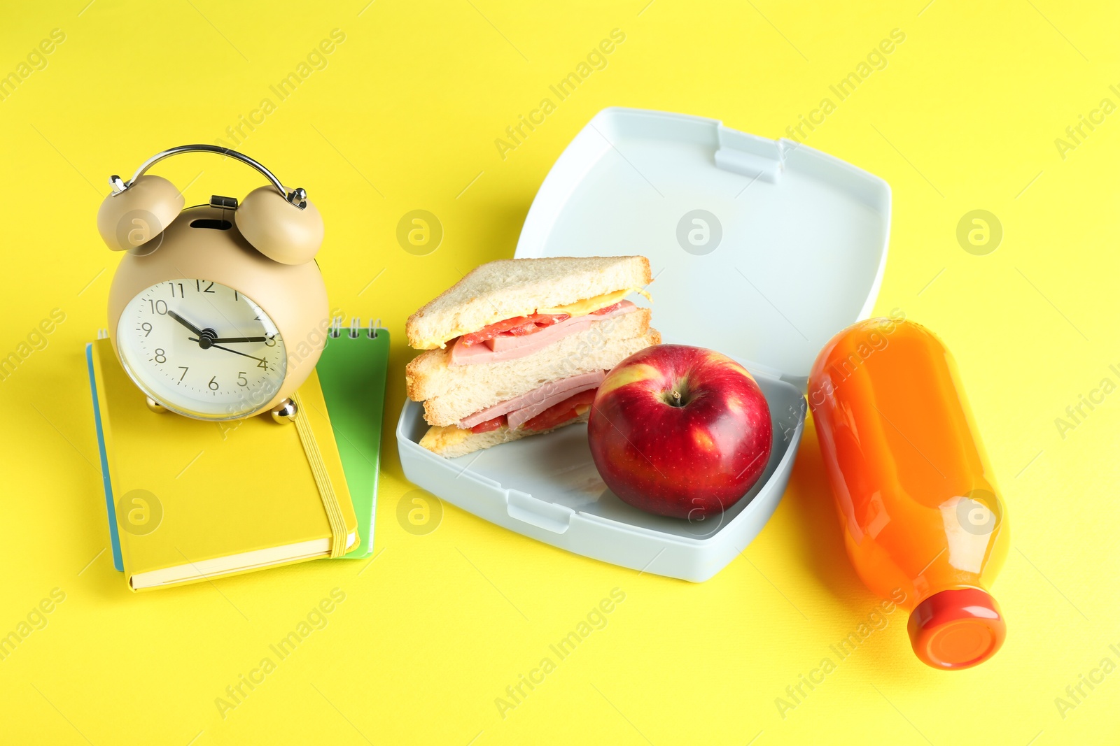 Photo of Lunch box with snacks, bottle of juice, alarm clock and books on yellow background