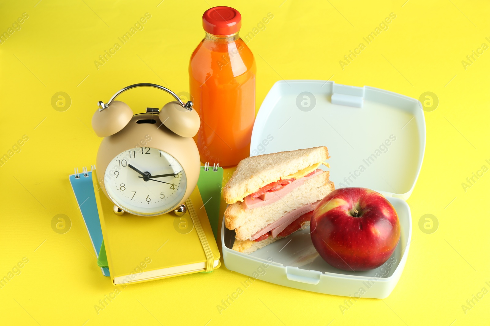 Photo of Lunch box with snacks, bottle of juice, alarm clock and books on yellow background