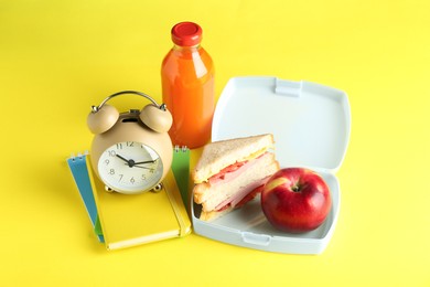 Photo of Lunch box with snacks, bottle of juice, alarm clock and books on yellow background