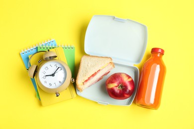 Photo of Lunch box with snacks, bottle of juice, alarm clock and books on yellow background, flat lay