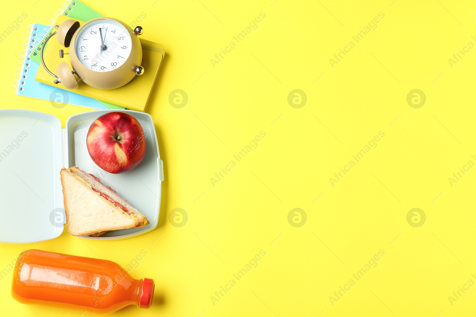 Photo of Lunch box with snacks, bottle of juice, alarm clock and books on yellow background, flat lay. Space for text