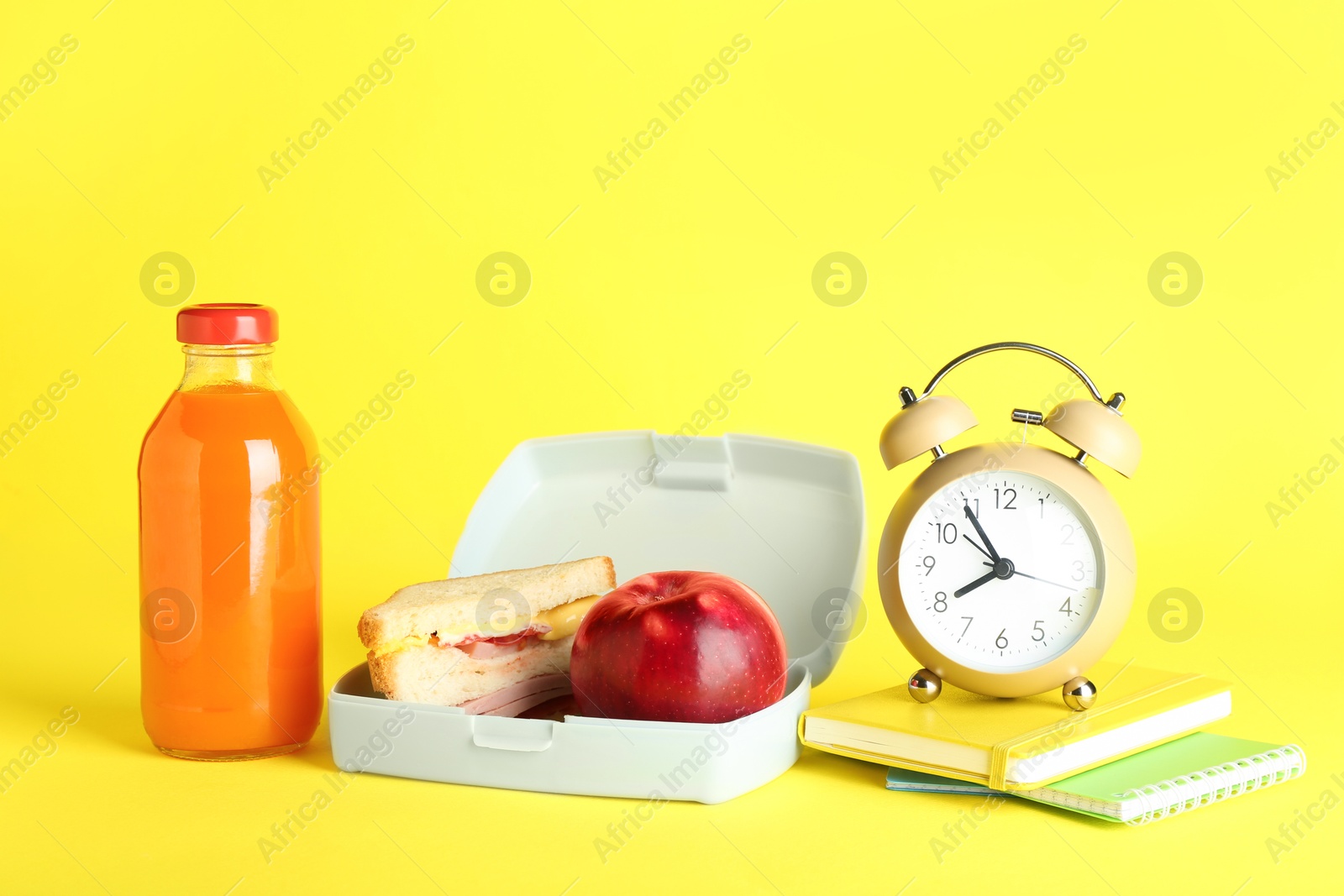 Photo of Lunch box with snacks, bottle of juice, alarm clock and books on yellow background