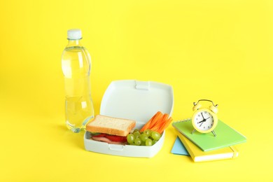 Photo of Lunch box with snacks, bottle of water, alarm clock and books on yellow background