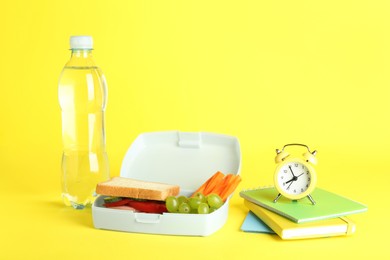 Photo of Lunch box with snacks, bottle of water, alarm clock and books on yellow background