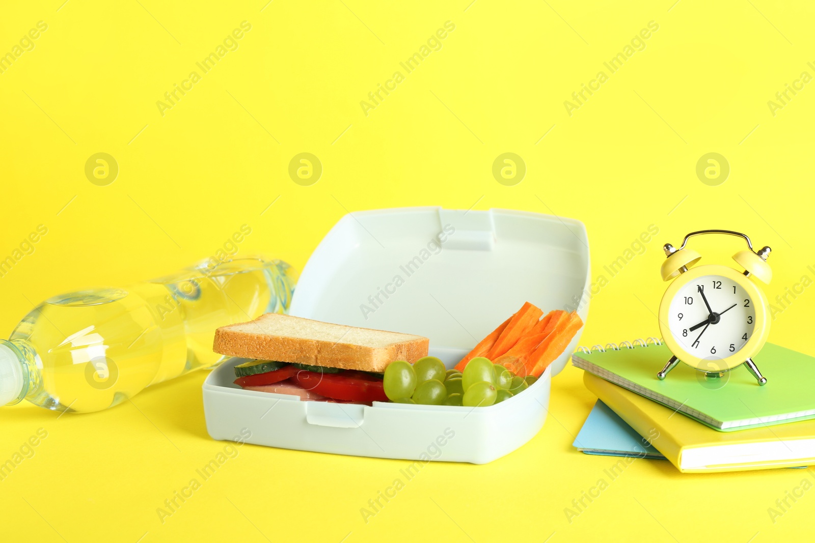 Photo of Lunch box with snacks, bottle of water, alarm clock and books on yellow background