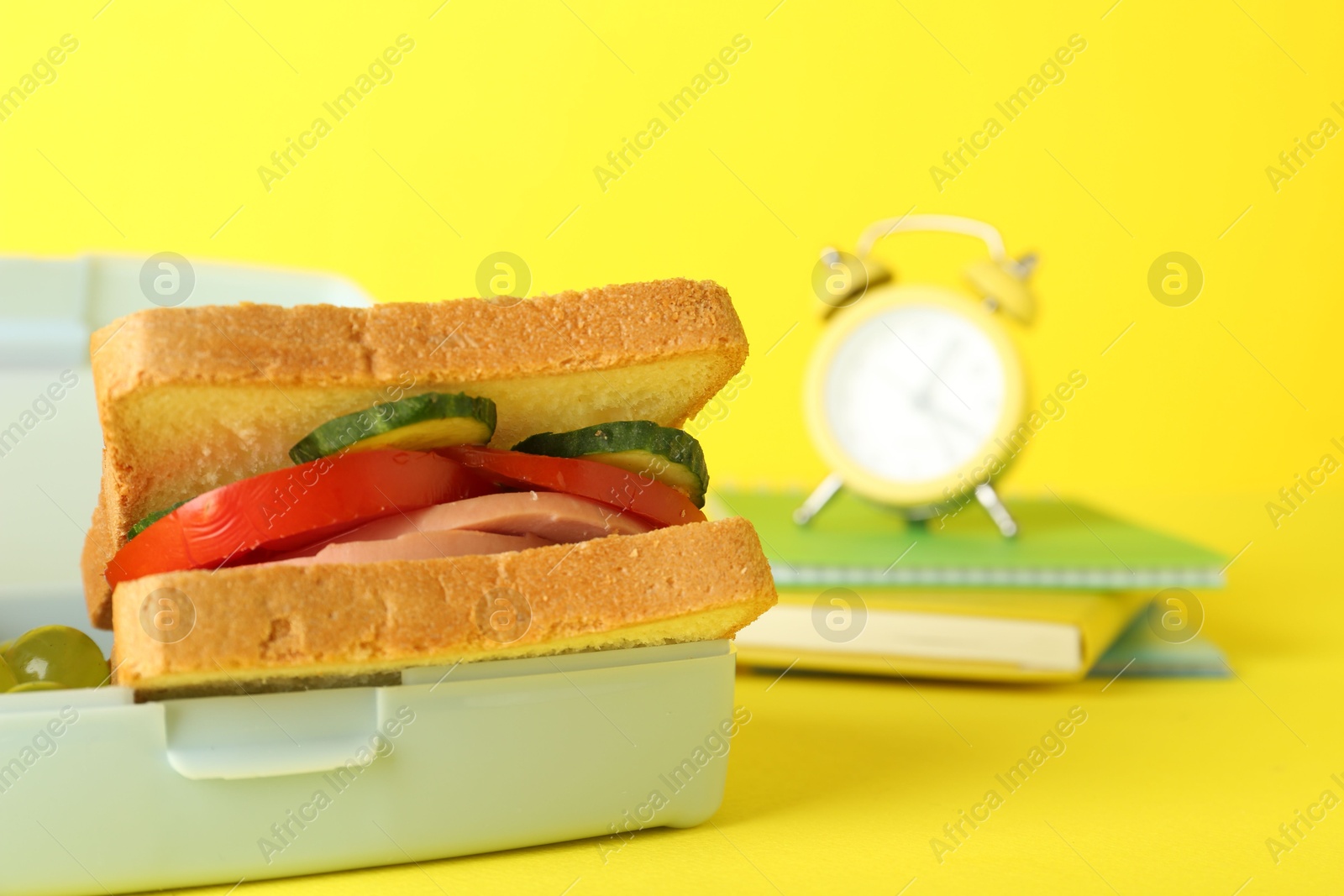 Photo of Lunch box with snacks, alarm clock and books on yellow background, closeup