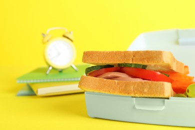 Photo of Lunch box with snacks, alarm clock and books on yellow background, closeup