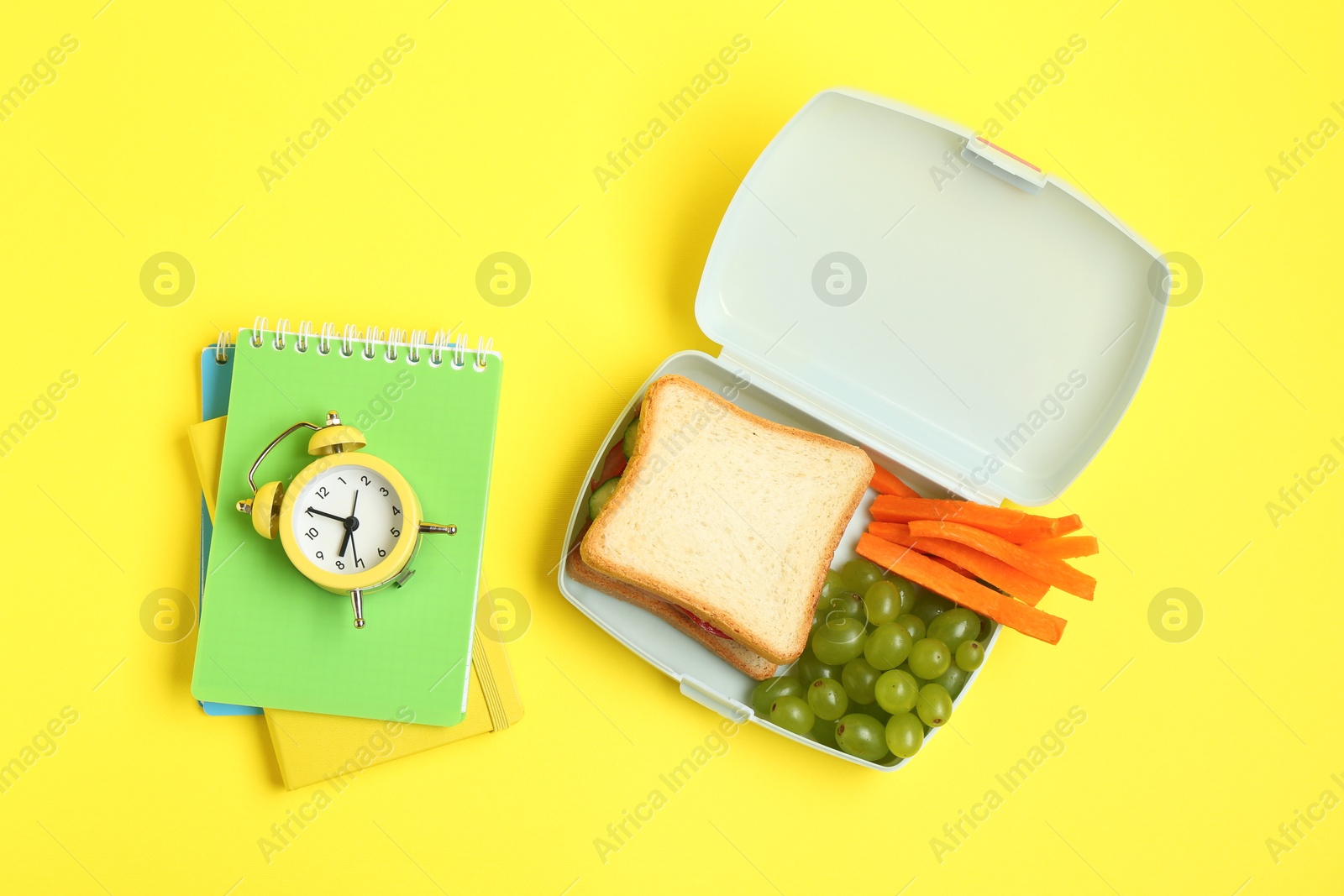 Photo of Lunch box with snacks, alarm clock and books on yellow background, flat lay