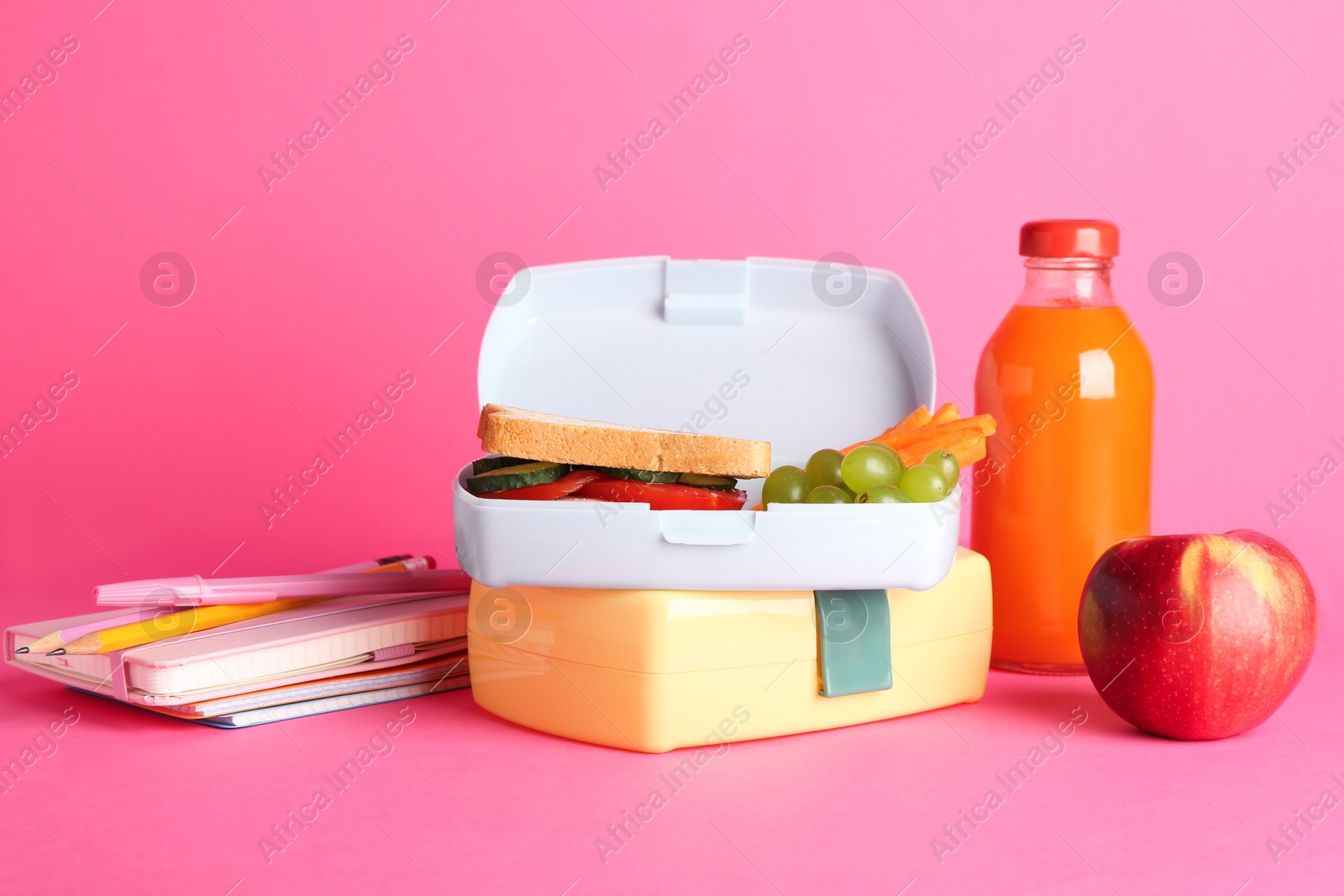 Photo of Lunch boxes with snacks, bottle of juice, alarm clock and stationery on pink background