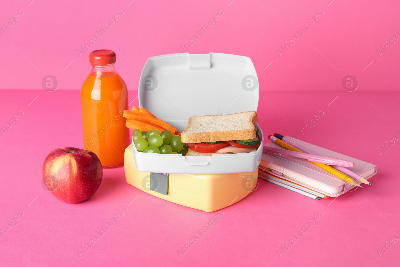 Photo of Lunch boxes with snacks, bottle of juice, alarm clock and stationery on pink background