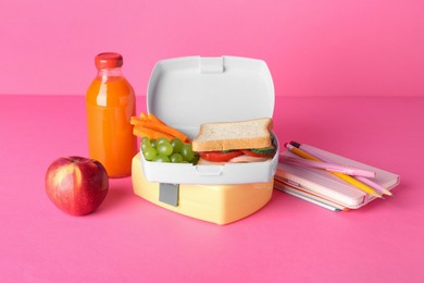 Photo of Lunch boxes with snacks, bottle of juice, alarm clock and stationery on pink background