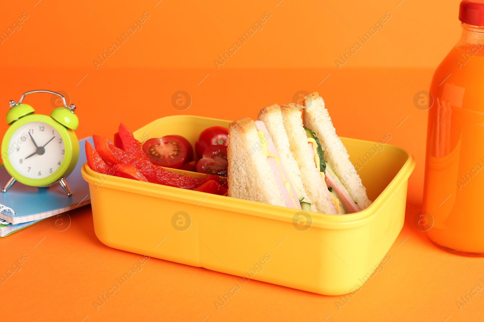 Photo of Lunch box with snacks, bottle of juice, alarm clock and notebooks on orange background, closeup