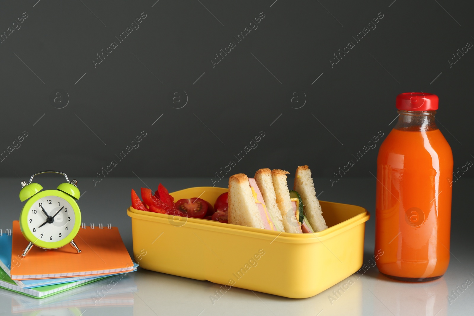 Photo of Lunch box with snacks, bottle of juice, alarm clock and stationery on light table