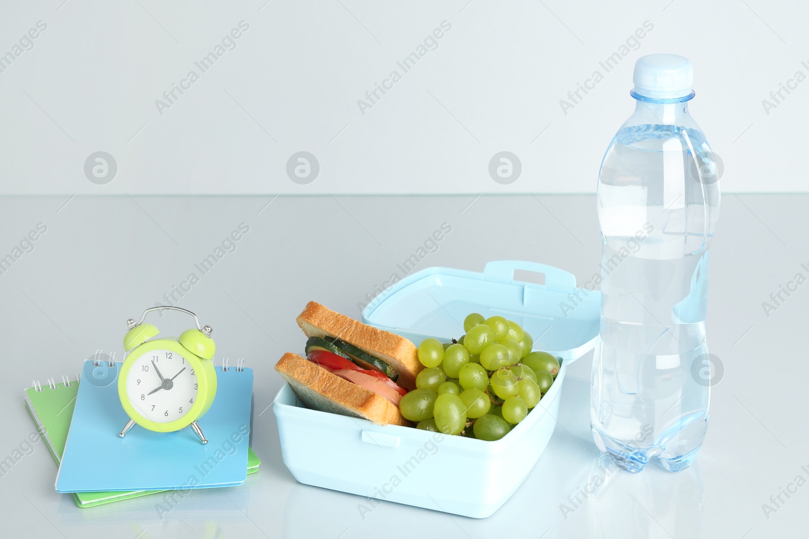 Photo of Lunch box with snacks, bottle of water, alarm clock and stationery on white table