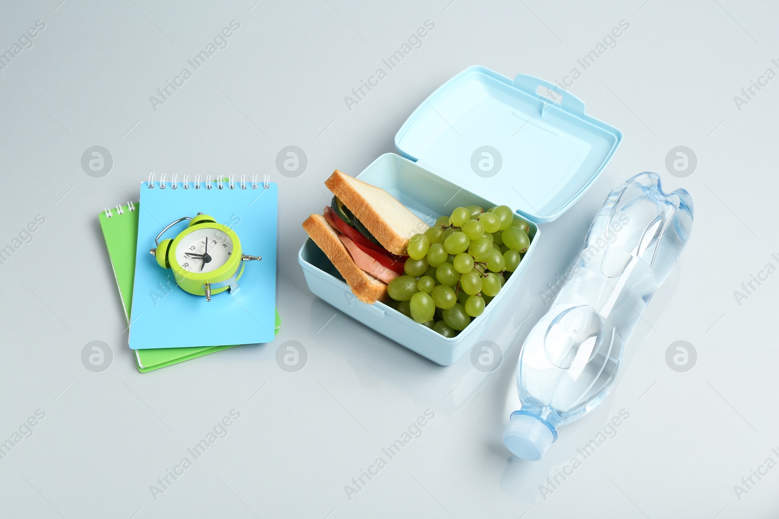 Photo of Lunch box with snacks, bottle of water, alarm clock and stationery on white table