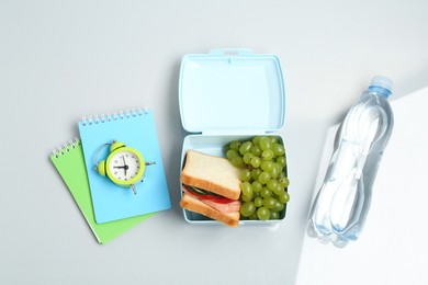 Photo of Lunch box with snacks, bottle of water, alarm clock and stationery on white background, flat lay