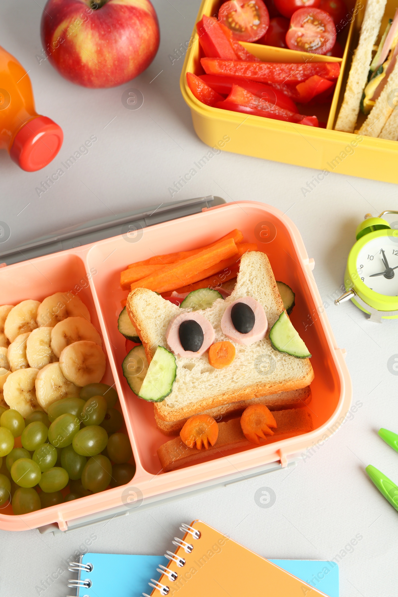 Photo of Lunch boxes with snacks, bottle of juice, alarm clock and stationery on white table