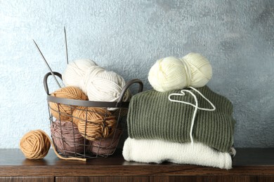 Photo of Basket with colorful yarns, knitting needles and stack of sweaters on wooden shelf indoors