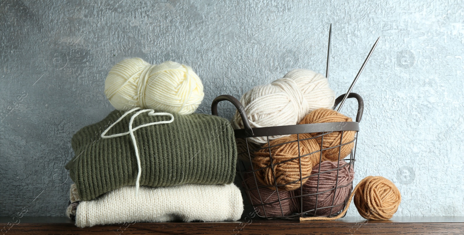 Photo of Basket with colorful yarns, knitting needles and stack of sweaters on wooden shelf indoors