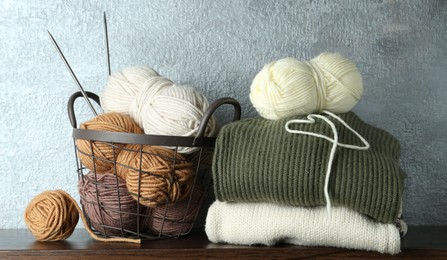 Photo of Basket with colorful yarns, knitting needles and stack of sweaters on wooden shelf indoors