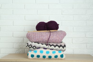 Skein of yarn, knitting needles and sweaters on wooden shelf near white brick wall