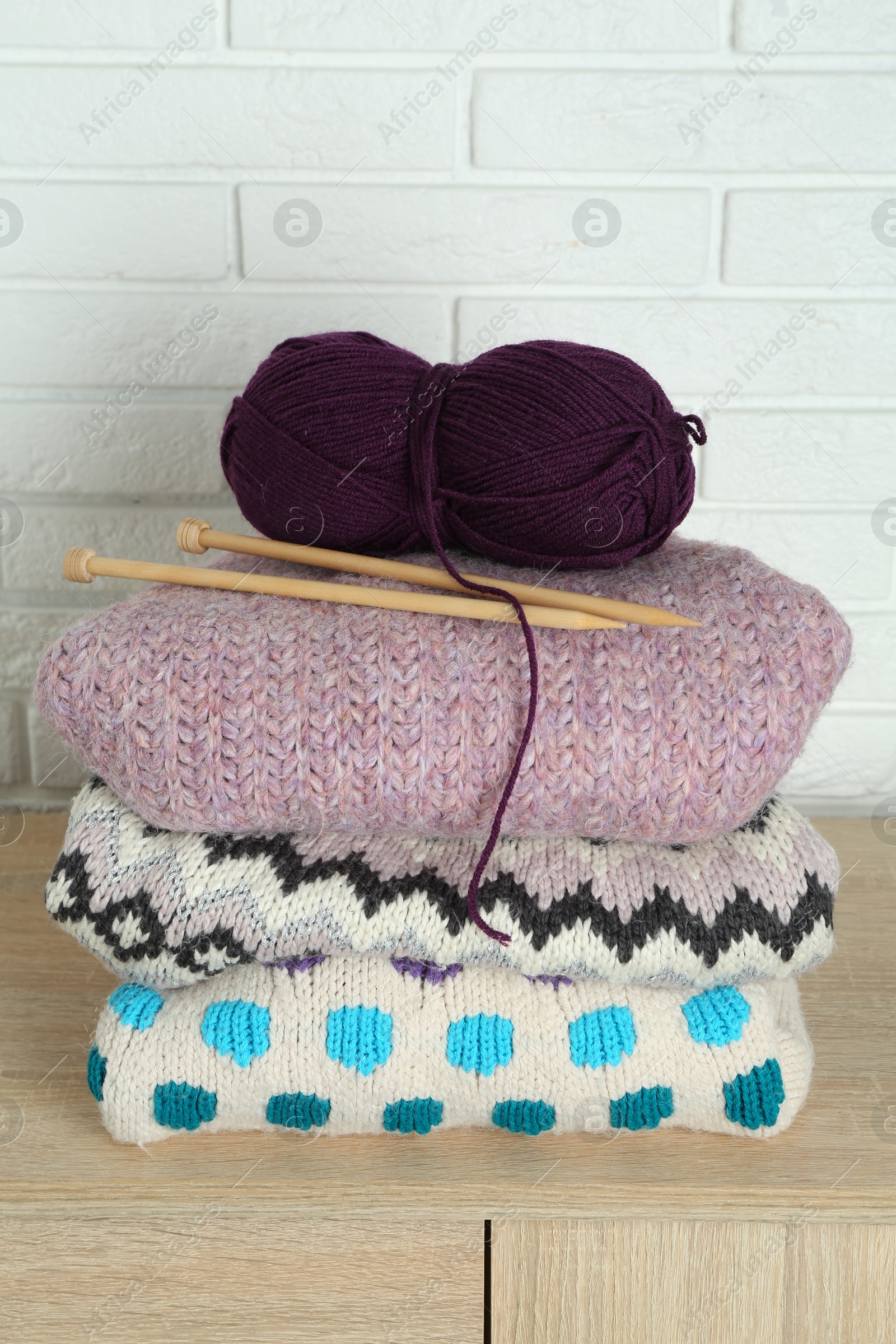 Photo of Skein of yarn, knitting needles and sweaters on wooden chest of drawers near white brick wall