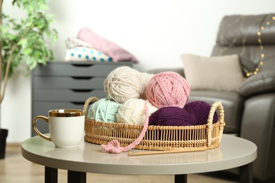 Photo of Wicker basket with colorful yarns, crochet pattern, hook and cup on coffee table indoors