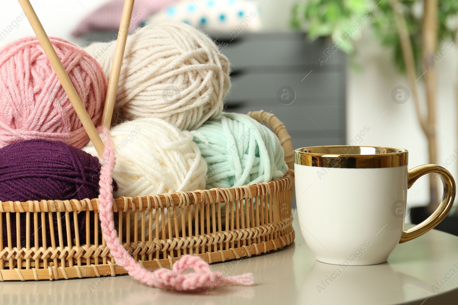 Photo of Wicker basket with colorful yarns, crochet pattern and cup on coffee table indoors, closeup