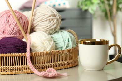 Photo of Wicker basket with colorful yarns, crochet pattern and cup on coffee table indoors, closeup