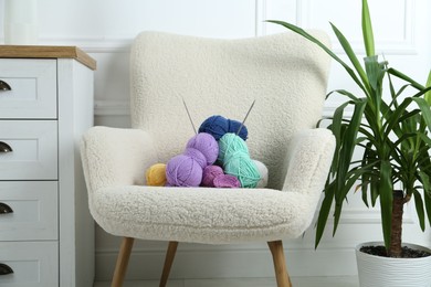 Photo of Armchair with pile of colorful yarns, crochet pattern and knitting needles between houseplant and chest of drawers indoors