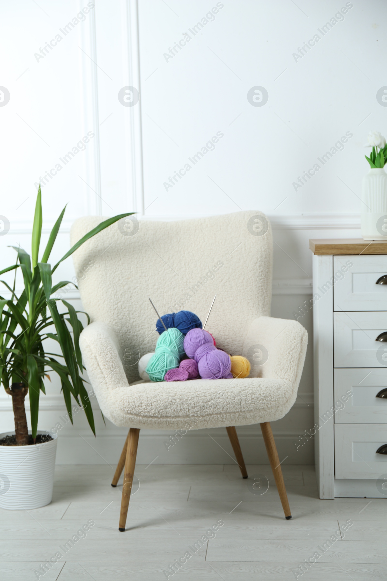 Photo of Armchair with pile of colorful yarns, crochet pattern and knitting needles between houseplant and chest of drawers indoors