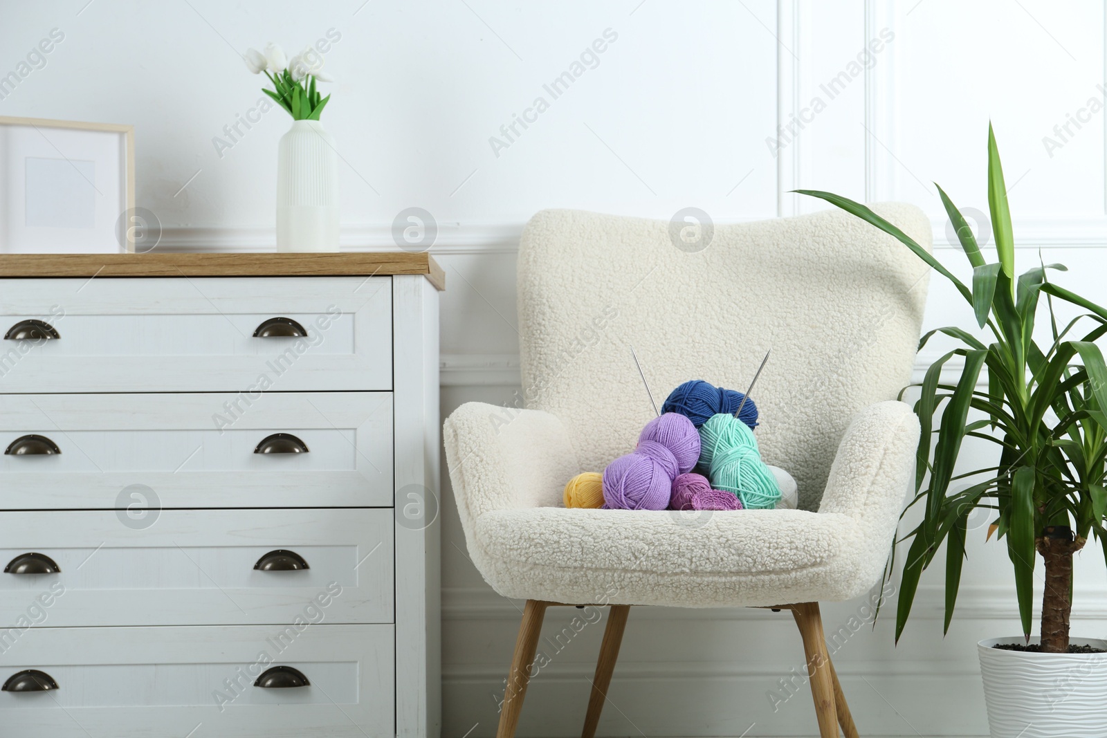 Photo of Armchair with pile of colorful yarns, crochet pattern and knitting needles between houseplant and chest of drawers indoors