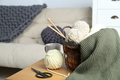 Basket with yarns, knitting needles, scissors and sweater on wooden table indoors