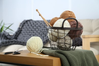 Photo of Basket with yarns, knitting needles, scissors and sweater on wooden table indoors