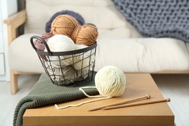 Photo of Basket with yarns, knitting needles and sweater on coffee table indoors