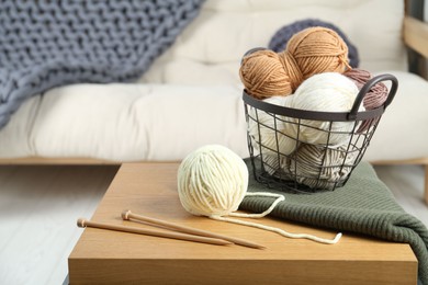 Photo of Basket with yarns, knitting needles and sweater on coffee table indoors