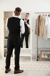 Handsome man adjusting bow tie near mirror indoors