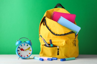 Photo of Backpack with different school stationery and alarm clock on white table against green background