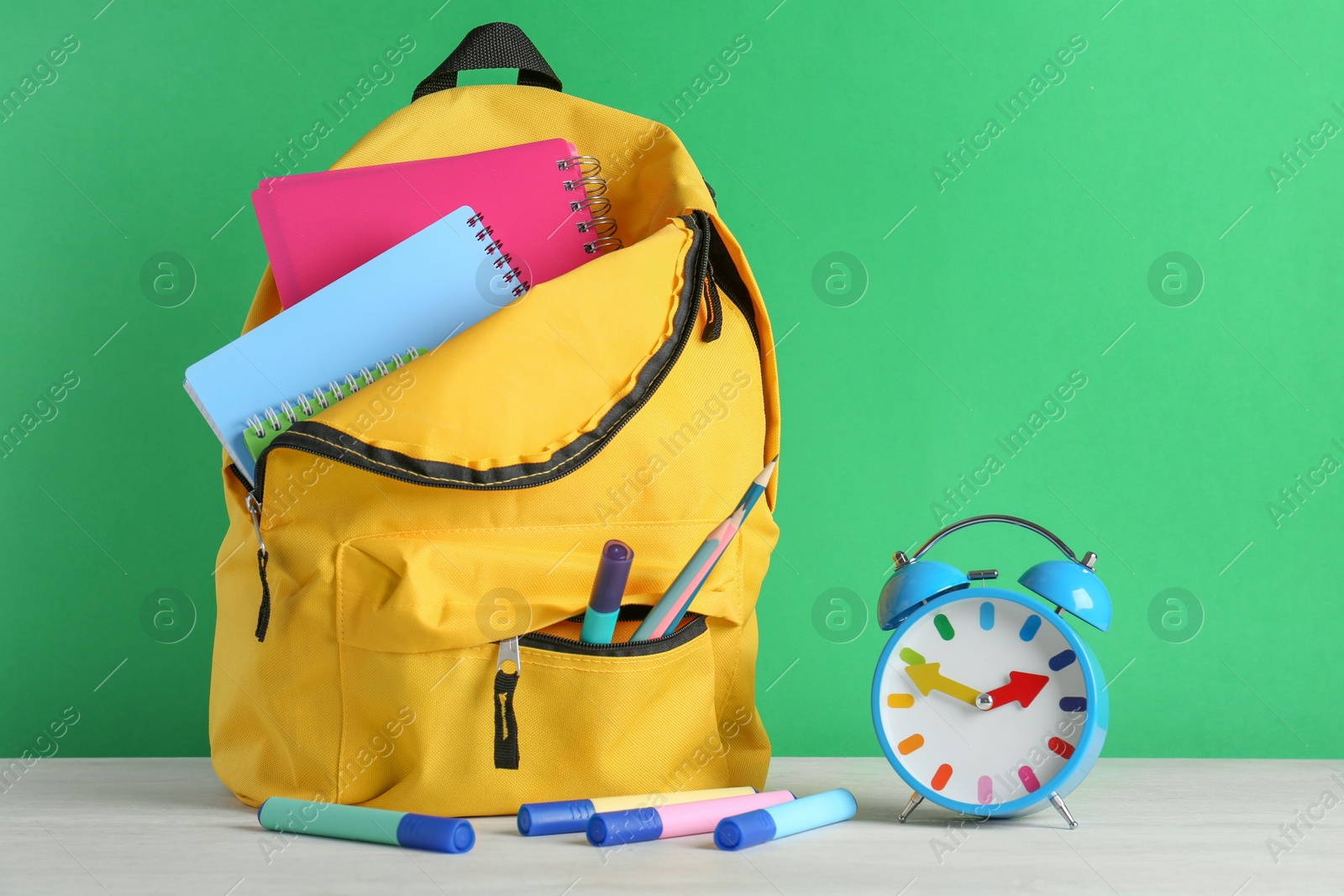 Photo of Backpack with different school stationery and alarm clock on white table against green background