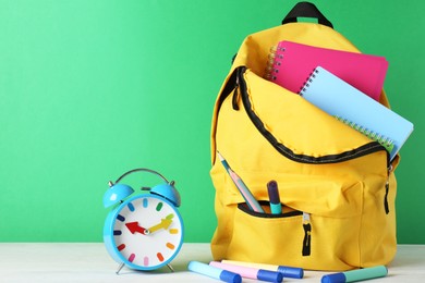 Photo of Backpack with different school stationery and alarm clock on white table against green background