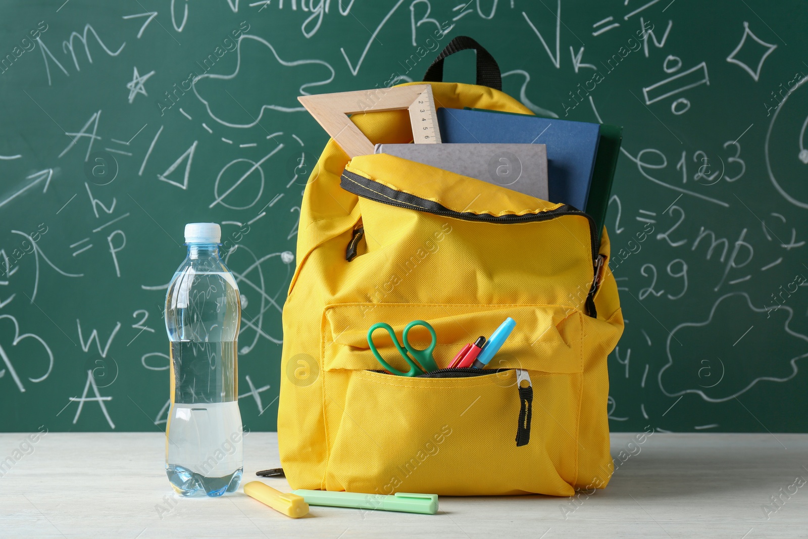 Photo of Backpack with different school stationery and bottle of water on white table against green chalkboard