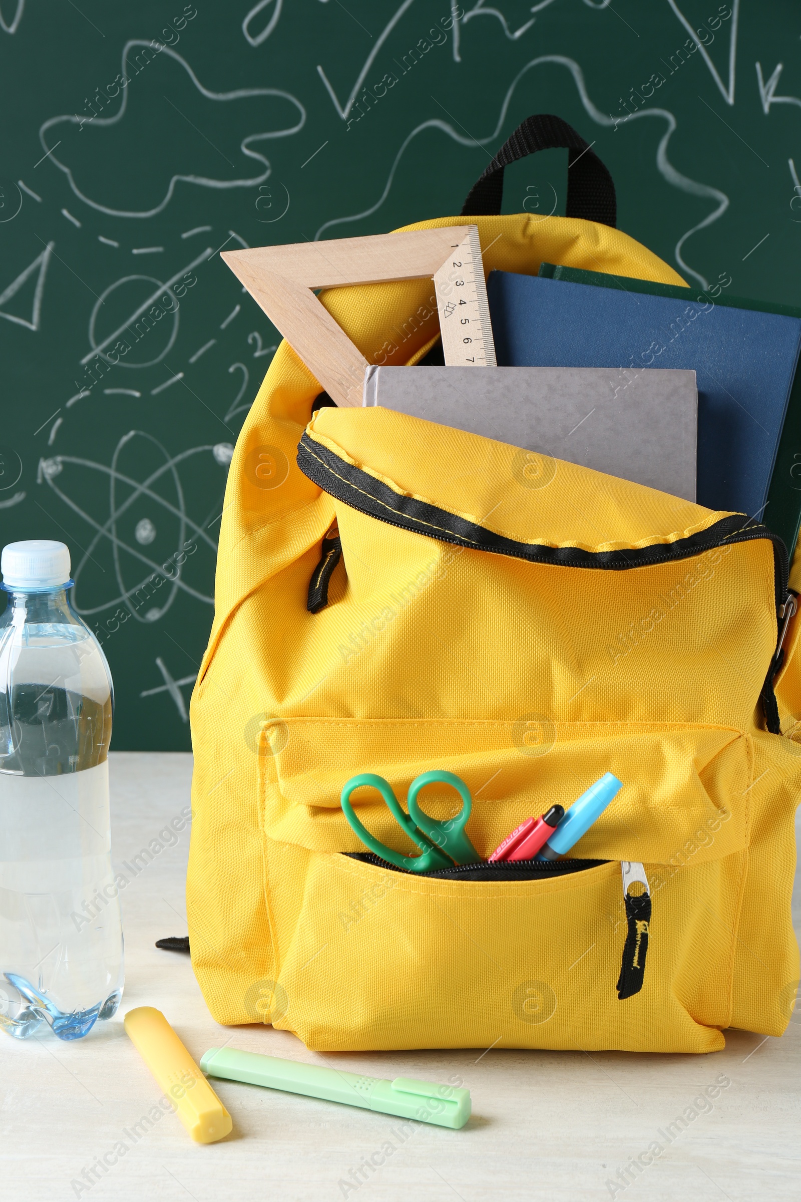 Photo of Backpack with different school stationery and bottle of water on white table against green chalkboard